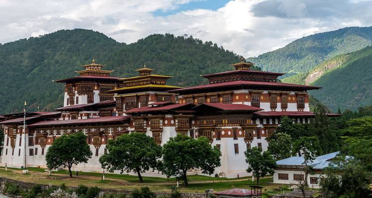 Majestic riverside dzong with ornate wooden windows framed by lush mountains and cloudy sky.