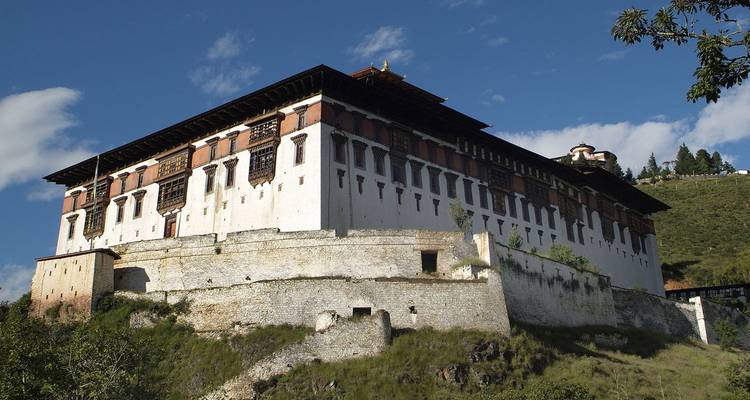 Large white-walled fortress dzong atop a grassy hill under deep blue sky with scattered clouds.