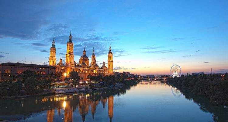 La Basílica de Nuestra Señora del Pilar iluminada se refleja en el río Ebro a la hora azul en Zaragoza.
