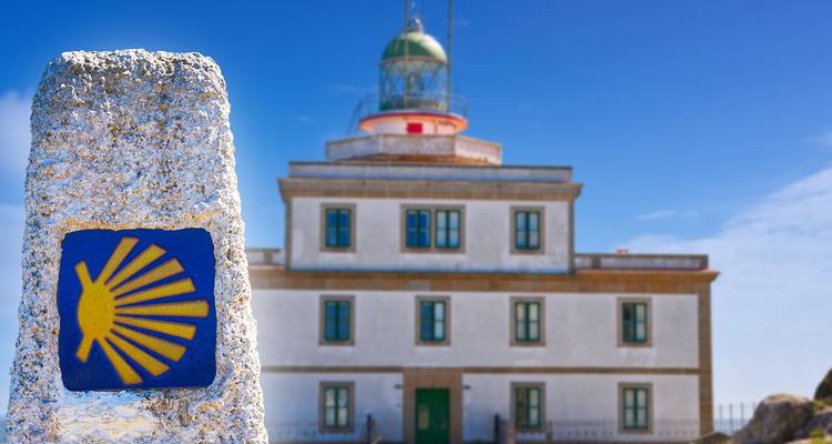 Marcador de piedra del Camino con concha de vieira amarilla frente a un faro blanco bajo cielo azul
