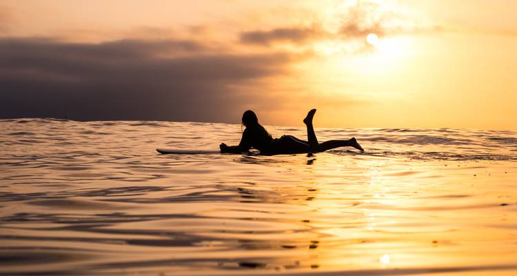 Surfista silueteado acostado en una tabla esperando olas durante una puesta de sol brillante