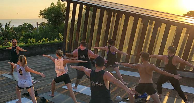 Clase de yoga al aire libre fluyendo a través de posturas de guerrero en una terraza de madera al amanecer junto al mar