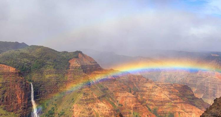 Un arc-en-ciel s'étend au-dessus de falaises rouges escarpées et d'une cascade lointaine dans le canyon de Waimea sous un ciel brumeux.