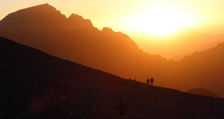 Des randonneurs en silhouette se tenant par la main pendant qu'ils gravissent une pente d'éboulis escarpée face à un lever de soleil dramatique dans le Haut Atlas.