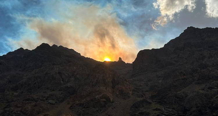 Le soleil se profile entre les crêtes montagneuses escarpées, projetant des nuages fumeux qui rougeoient contre un ciel bleu.