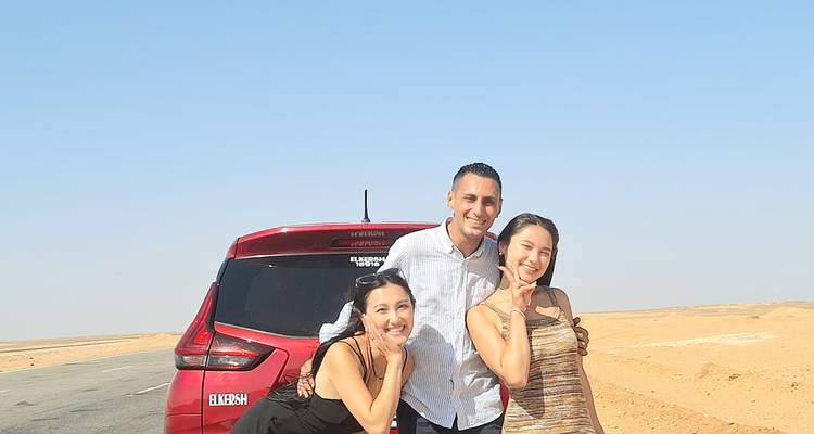 Deux femmes et un homme posent en souriant à côté d'une voiture rouge sur une route désertique désolée.