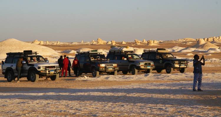 Flotte de jeeps et voyageurs rassemblés sur un terrain désertique blanc et plat éclairé par la chaleur du soleil en fin d'après-midi.
