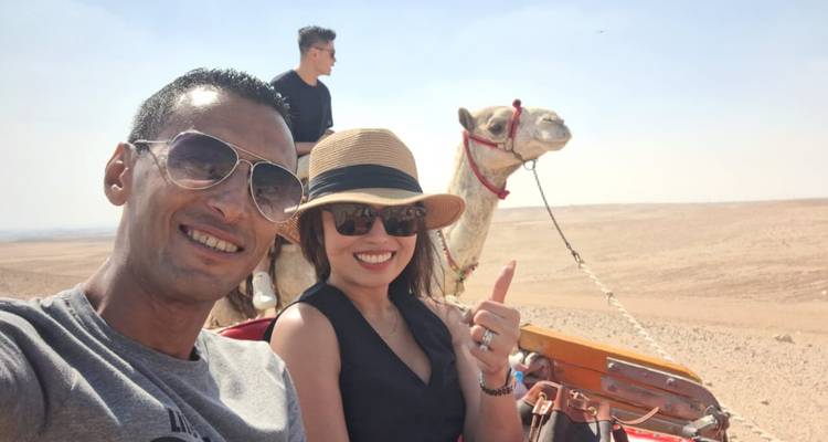 Un couple prend un selfie à dos de chameau tandis qu'un guide accompagne leur monture à travers le désert près des pyramides.