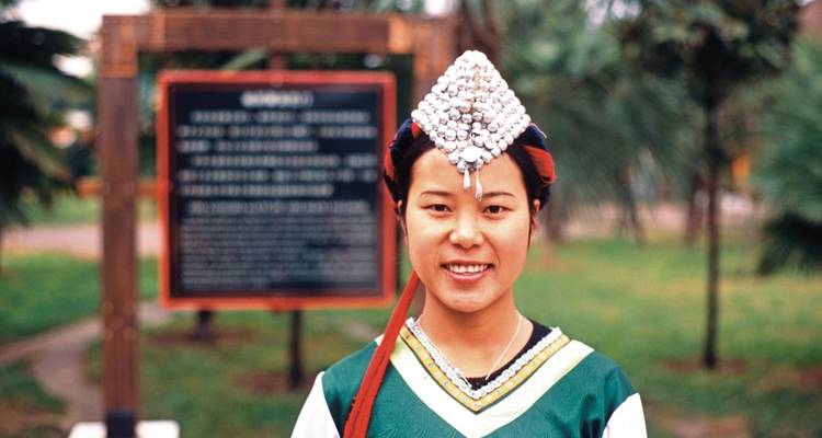 Una mujer sonriente con vestimenta tradicional, con vegetación en el fondo.