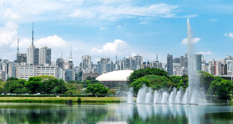 Stadssilhouet van São Paulo weerspiegeld in rustig meer met fontijnen die water spuiten.