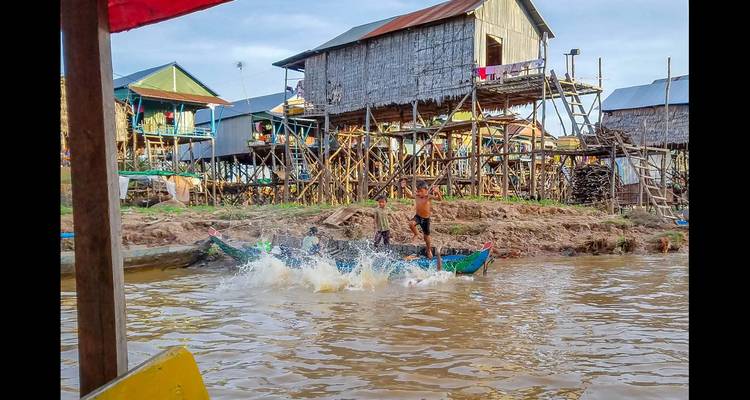 Des enfants jouent dans une petite barque devant des maisons sur pilotis au bord de l'eau.