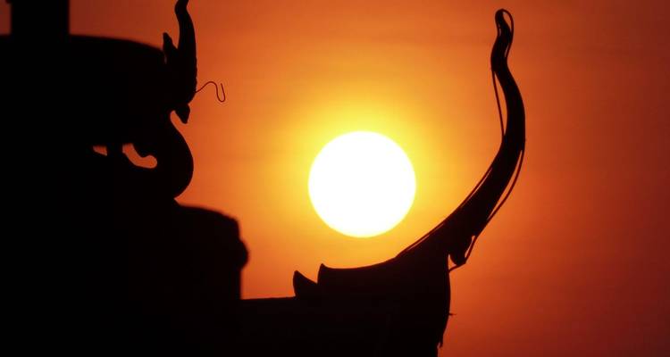 Un sol anaranjado ardiente se pone detrás de la elegante silueta curva del ornamento del techo de un templo.