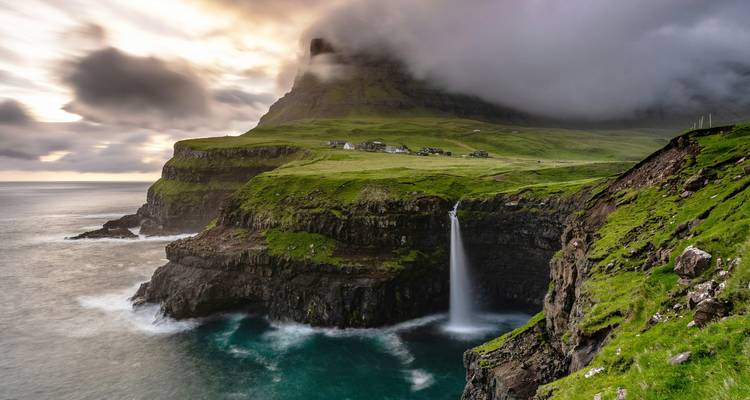 Waterfall flowing into the ocean beneath steep cliffs with distant houses.