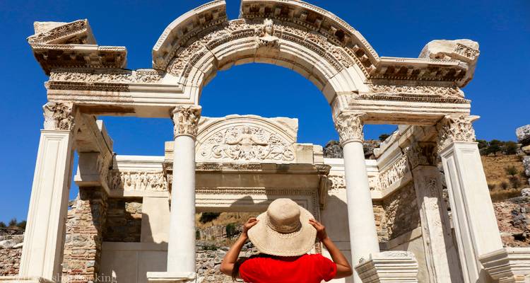 Viajero con sombrero de paja mira hacia arriba al arco ornamentado del Templo de Adriano en Éfeso bajo un cielo azul claro.