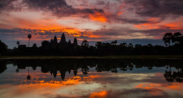 Silueta de Angkor Wat reflejada en un estanque quieto bajo nubes anaranjadas ardientes del amanecer.