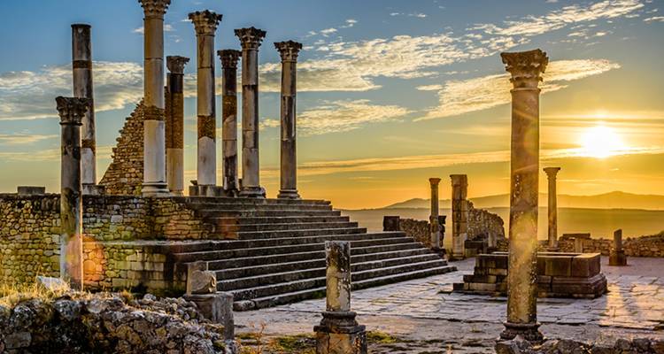 Ruines romaines antiques avec des colonnes et le soleil se couchant en arrière-plan.