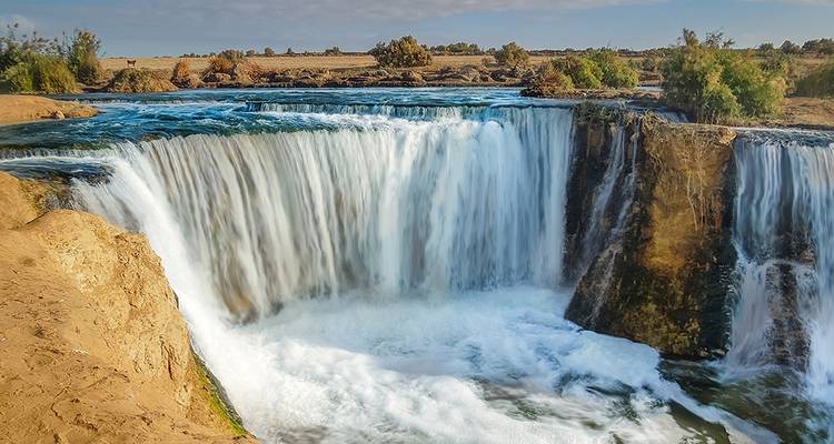 Ein Wasserfall, der in einen Fluss hinabstürzt, mit einem klaren blauen Himmel.