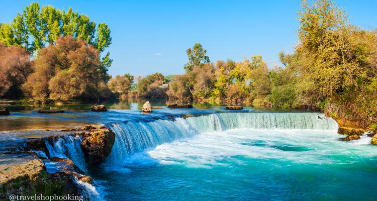 Amplio río turquesa cae en cascada sobre una catarata baja rodeada de árboles frondosos bajo un cielo despejado.