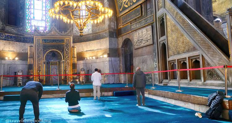 Besucher knien und beten in einer reich verzierten Moschee mit blauem Teppich und einem prächtigen Kronleuchter.