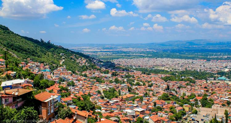 Panoramic vista over Bursa with red-roofed houses climbing a green hillside under a partly cloudy sky