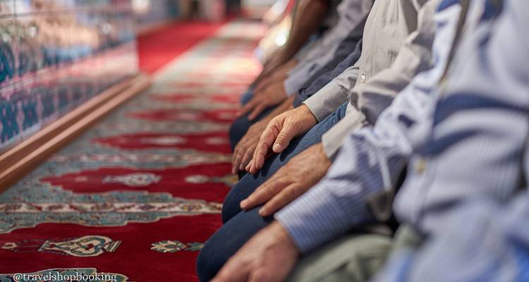 Row of worshippers kneel on patterned carpet inside a mosque with focus on their resting hands