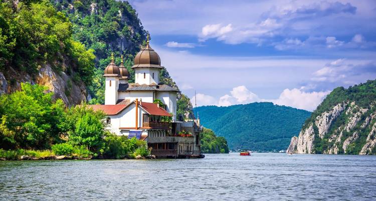 Een kerk aan de rivieroever met heuvels en een bewolkte hemel op de achtergrond.
