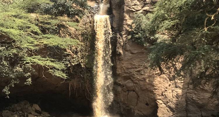 Ein kleiner Wasserfall, umgeben von felsigem Gelände und Grün.