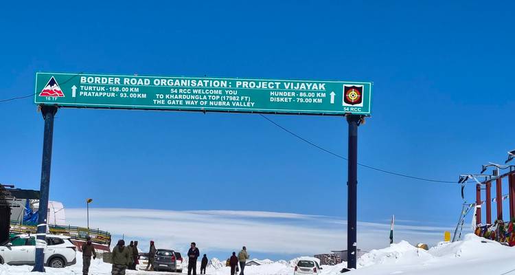 Col de montagne enneigé avec panneau vert de l'Organisation des routes frontalières, voitures garées et promeneurs sous un ciel bleu dégagé