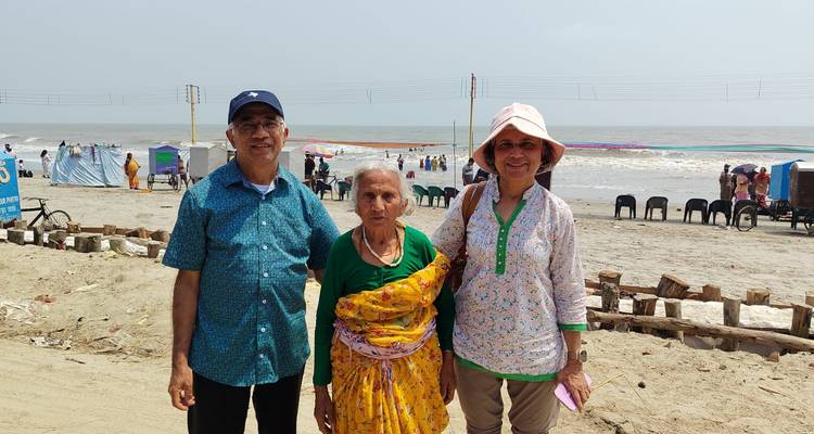 An elderly woman and two relatives pose on a sandy beach with gentle surf and beachgoers in the background.
