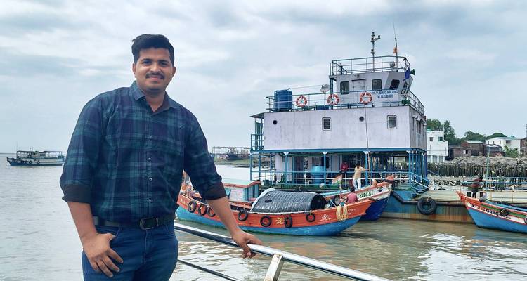 Smiling man posing on a boat deck with colorful fishing boats and a gray sky behind.