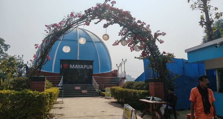 Un portal floral arqueado con un letrero de 'I ❤️ MAYAPUR' enmarca un edificio de cúpula azul bajo la luz solar brumosa.