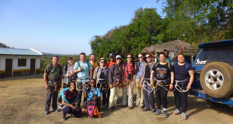 Un gran grupo de aventureros con arneses de escalada posando juntos en un campamento polvoriento al lado de un 4x4.