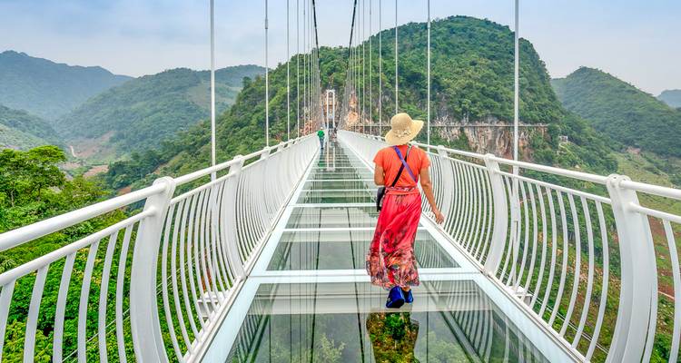Eine Frau mit Hut, die über eine Brücke mit Glasboden geht, mit üppigen grünen Bergen im Hintergrund.