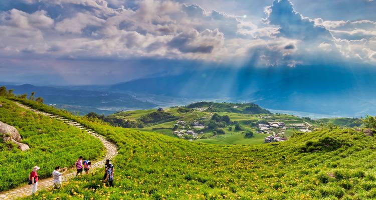 Zonnestralen breken door dramatische wolken boven glooiende groene heuvels bezaaid met gele bloemen and dorpen