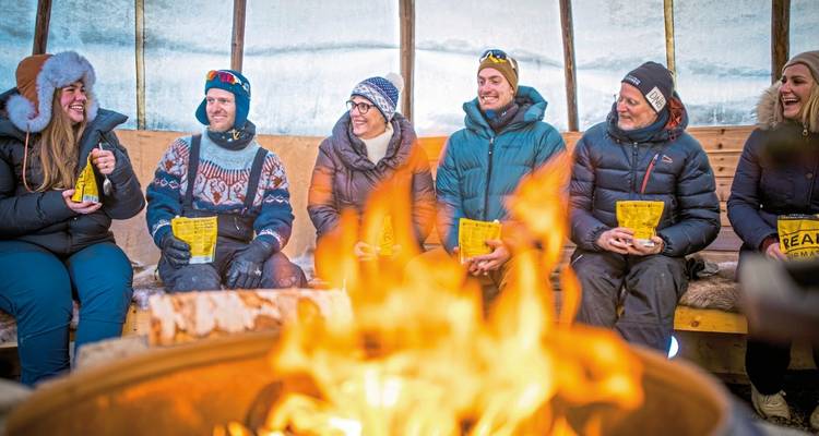 Groupe de personnes dans une cabane chaleureuse partageant une boisson chaude près du feu.