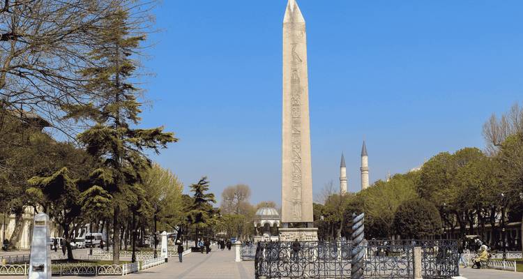 Steinobelisk, der im historischen Hippodrom von Istanbul steht, flankiert von Bäumen und Minaretten unter einem klaren blauen Himmel.