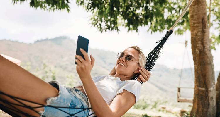 Femme se détendant dans un hamac, utilisant un smartphone avec vue sur la montagne.