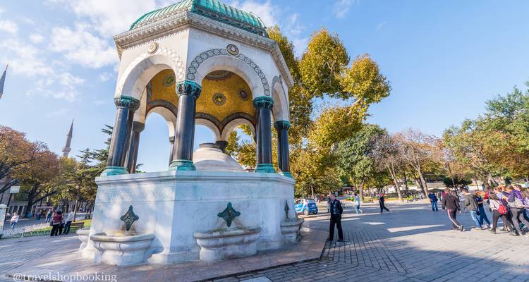 Seitliche Perspektive des Deutschen Brunnens in einer sonnigen Parkanlage mit Herbstlaub und Besuchern.