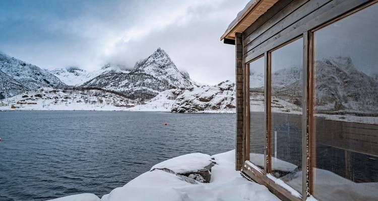 Chalet enneigé au bord d'un lac avec vue sur les montagnes.