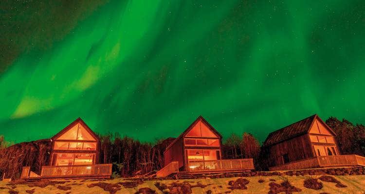 Aurores boréales au-dessus de maisons illuminées dans un environnement enneigé.
