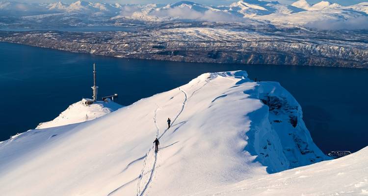Sommet de montagne enneigé avec traces de ski et océan au loin.