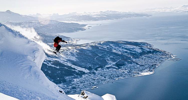 Skieur sautant d'une crête enneigée avec une vue panoramique sur un fjord.