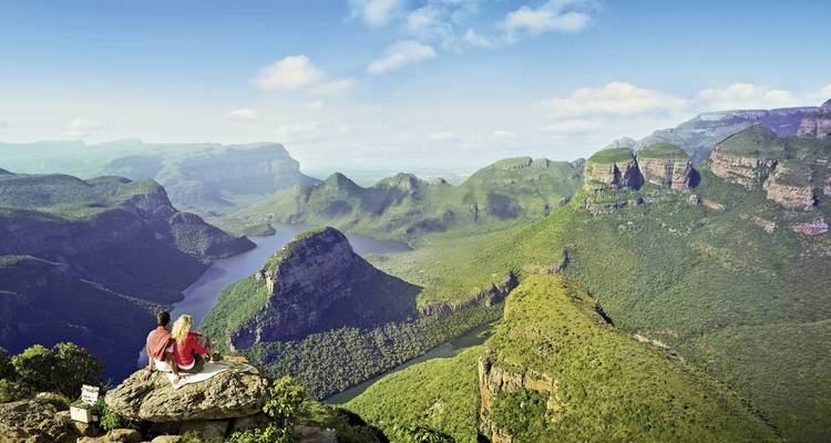 Dos personas sentadas en el borde de un acantilado contemplando un vasto paisaje de cañón verde.