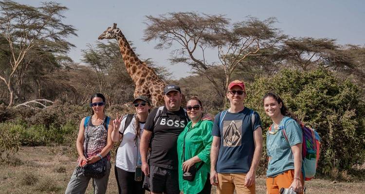 Gruppe von Touristen, die vor einer Giraffe in einer Savanne posieren.