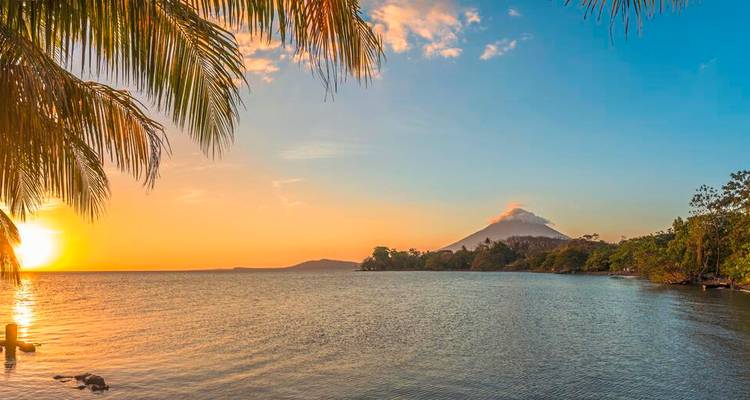 Coucher de soleil pittoresque sur un lac calme avec un volcan lointain et des frondes de palmier encadrant le ciel lumineux