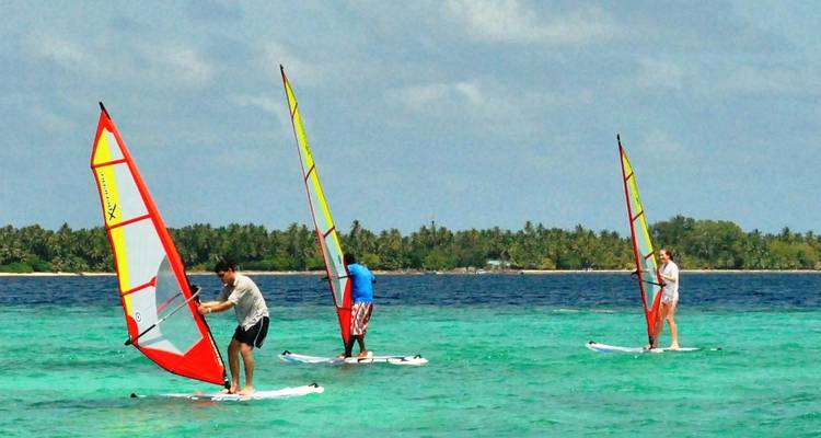 Drie windsurfers glijden over helder turquoise water nabij een met palmen omzoomd atol