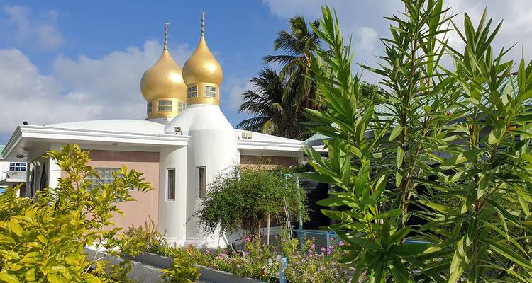Mosquée blanche avec deux dômes dorés en forme d'oignon entourée d'un feuillage de jardin luxuriant et de palmiers.