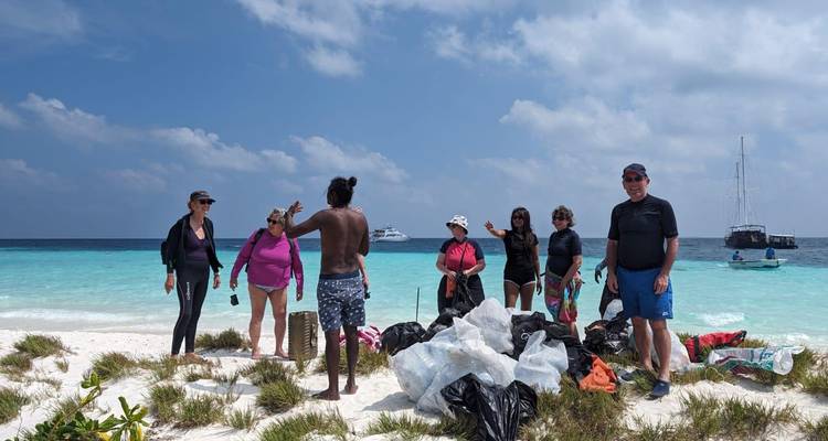 Gruppe von Freiwilligen, die auf einer unberührten Sandbank mit gesammelten Müllsäcken und türkisfarbenem Meer im Hintergrund stehen.