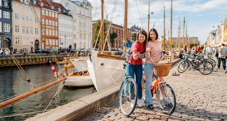 Zwei Menschen stehen mit Fahrrädern an einem Kanal, der von Booten und bunten Gebäuden gesäumt ist.
