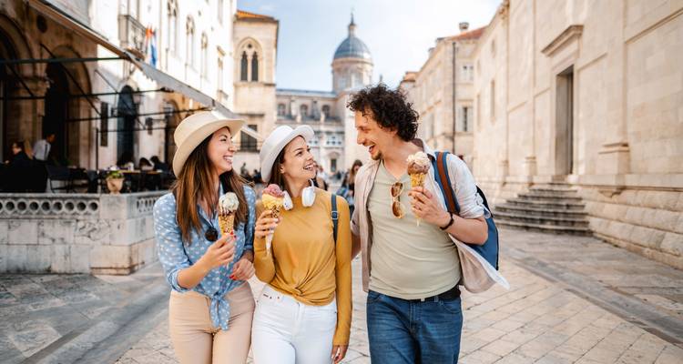 Drei Menschen gehen spazieren und genießen Eis in einer historischen Stadt mit einer Kathedrale im Hintergrund.